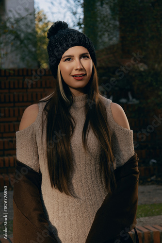 Mujer joven con gorro y bufanda de invierno posando casualmente en parque urbano soleado.