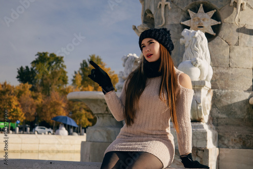 Mujer joven con gorro y bufanda de invierno posando casualmente en parque urbano soleado.
