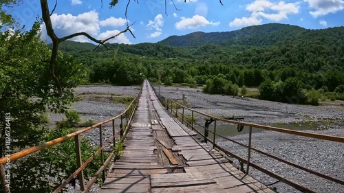 Worn, old wooden suspension bridge with missing planks and metal railings crosses wide riverbed. Fictional character walks across bridge. Natural landscape. Slow Motion Video, Not AI