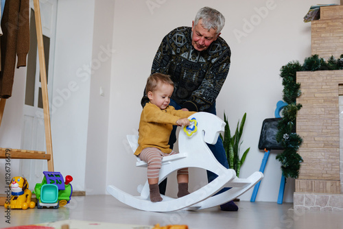 A joyful grandfather supports his young grandchild riding a white rocking horse inside a cozy room filled with toys. Warm moments are shared as they play