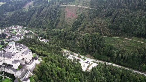 Drone flyby past Werfen Castle toward parking lot with forested mountain view
