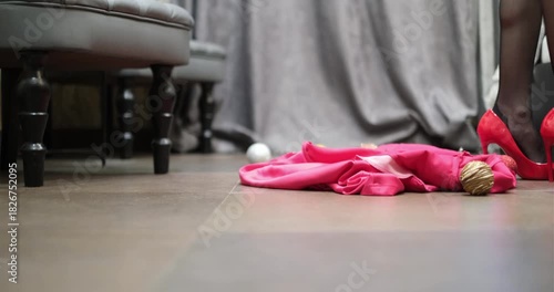 A woman takes off her dress, which falls to the floor and reveals female feet in stiletto heels
