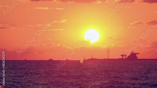 Number 1. Dramatic red-orange sunset, with the outline of coastline and ships and sailboats floating on water visible on horizon.