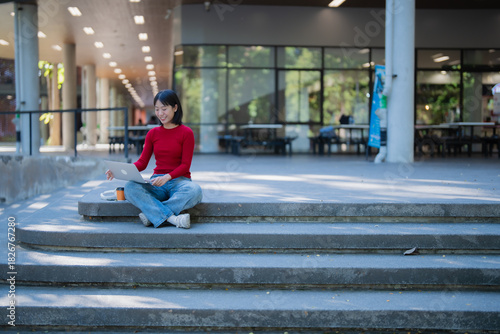 Asian female student studying on university campus stairs using laptop