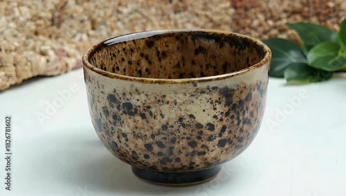 a high quality 4k scene showing a speckled ceramic mug standing empty on a clean white surface with a softly textured beige and brown background accented by fresh green plants creating a calm