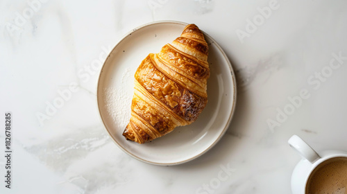 A golden brown, flaky croissant resting on a small, lightcolored ceramic plate, viewed from above on a white marble surface