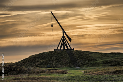 Fototapeta Naklejka Na Ścianę i Meble -  Silhouette of a Historic Trebuchet on a Hill at Sunset Over Open Grassland Landscape, Skagen, Denmark