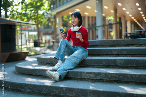 Asian woman smiling using phone on city stairs enjoying coffee
