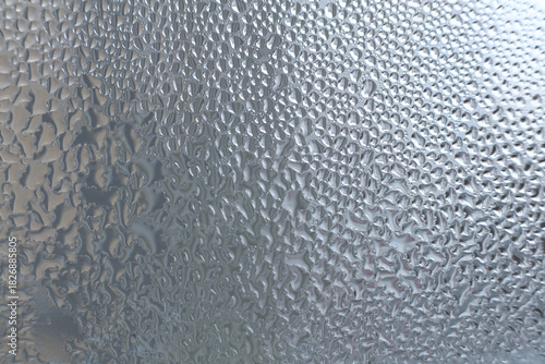 Macro close-up of condensation droplets on a smooth glass surface. A dense pattern of tiny water drops creates a textured abstract background with cool tones and soft natural light.