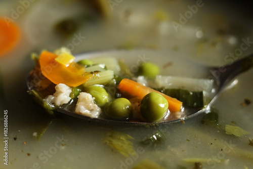 Macro close-up of a spoon filled with vegetables in clear broth. Visible peas, carrots, celery, and grains captured with shallow depth of field and warm natural light. Detailed food texture