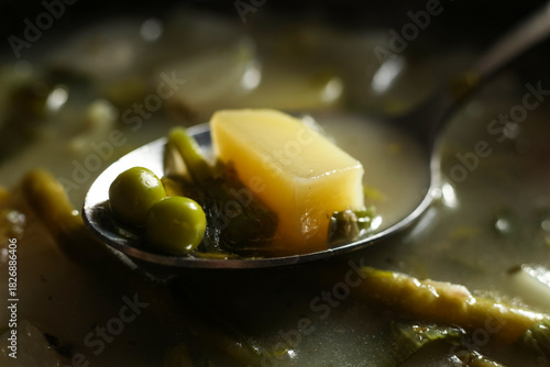 Macro close-up of a spoon filled with vegetables in clear broth. Visible peas, carrots, celery, and grains captured with shallow depth of field and warm natural light. Detailed food texture
