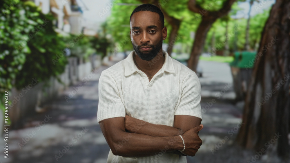 Fototapeta premium Man with arms crossed on a tree lined street looking downward in a white polo shirt wearing a beaded bracelet by a residential wall; contemplation.