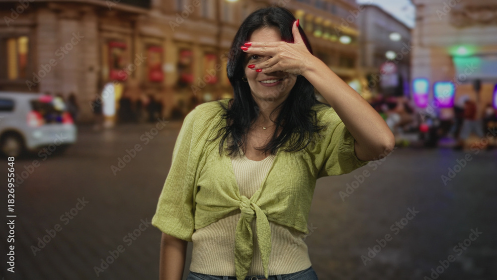 Fototapeta premium Woman covers eyes with a hand gesture in front of a lit building on street crowded with pedestrians; playfulness.