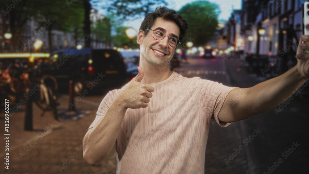 Fototapeta premium Man taking selfie with smartphone while giving thumbs up on a busy evening city street; playful confidence.
