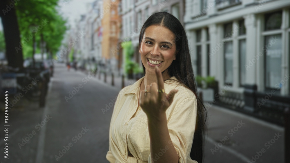Fototapeta premium Woman beckons with finger and smiles warmly on a cobbled city street lined with buildings and trees; invitation welcoming.