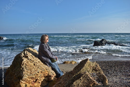 An elderly woman on the shore of a stormy sea in cool autumn weather.