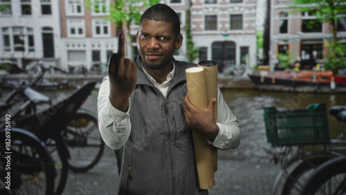 Photography Man holding rolled blueprints and raising middle finger, hand visible, on a canal street with bicycles and buildings; urban defiance