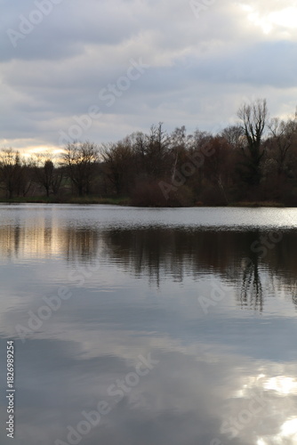 Photographie d’un lac paisible au crépuscule, avec les silhouettes d’arbres se reflétant sur l’eau calme. Une scène naturelle sereine idéale pour illustrer tranquillité, nature ou paysages d’extérieur