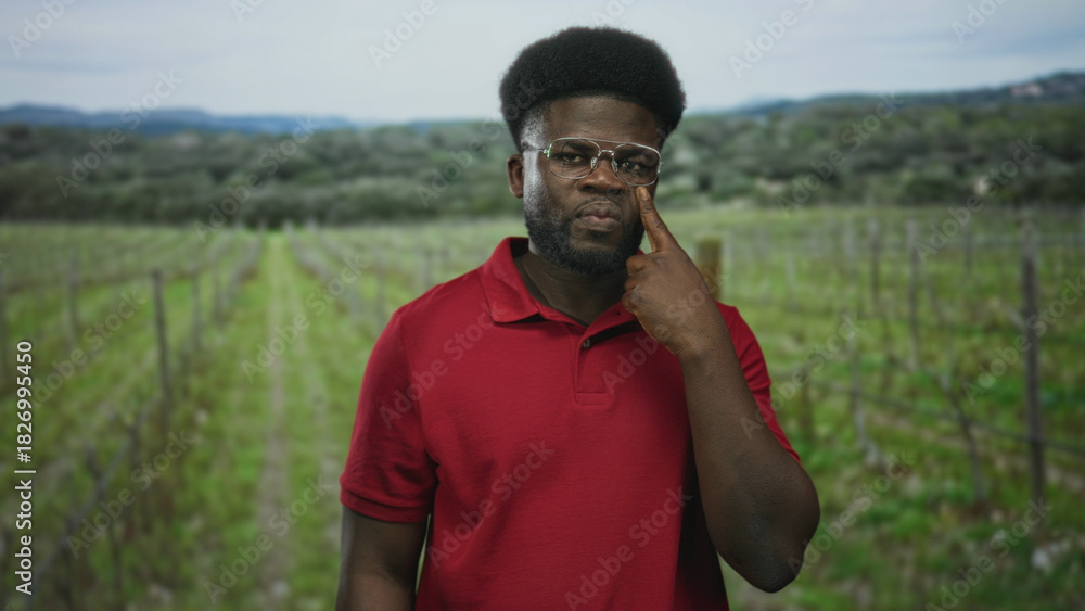 Fototapeta premium Man with glasses and beard, finger to cheek near eye in forest vineyard rows wearing red polo; pensive reflection.