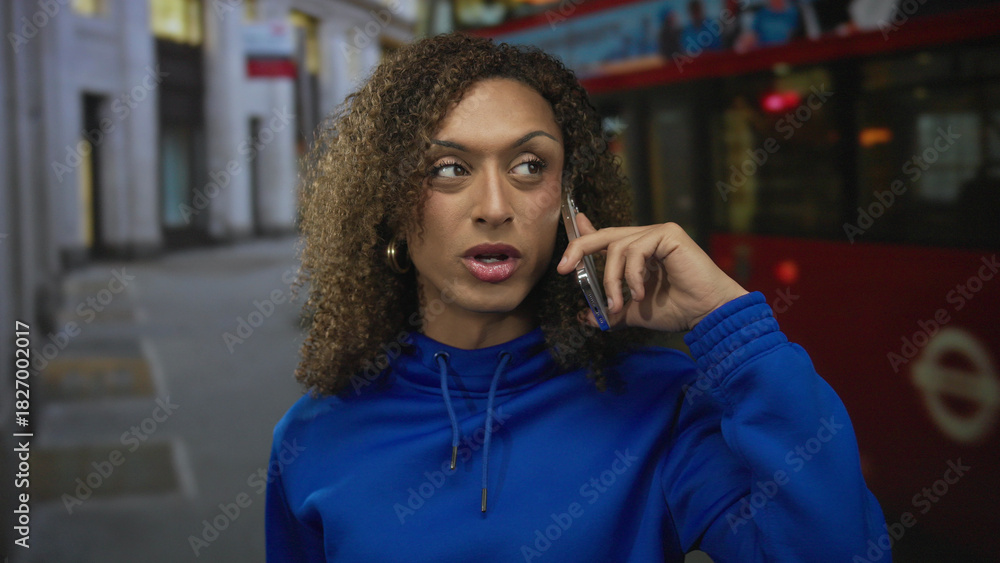 Fototapeta premium Woman wearing blue hoodie holding smartphone to ear on city street with red bus behind; isolation reflection.