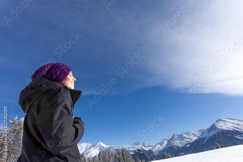 A person enjoys the warmth of the sun while standing in a snowy mountain landscape. With eyes closed and a peaceful smile, they embrace the crisp air and stunning alpine scenery