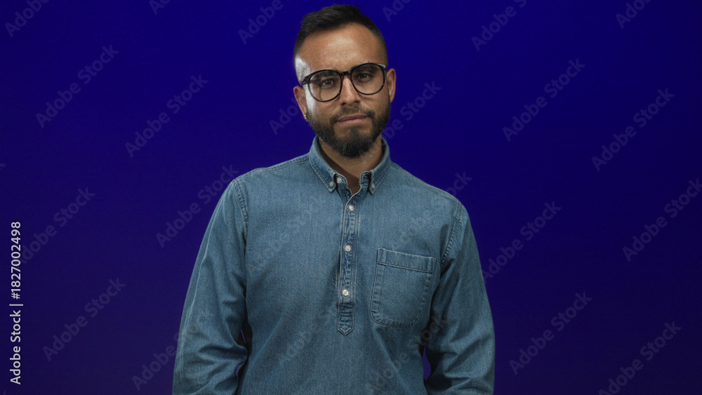 Obraz premium Young hispanic man wearing denim shirt with glasses flexes arm and shows bicep in blue studio; confidence strength.