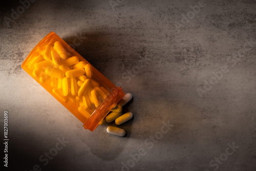 A dramatic, low-key photograph of capsules and pills spilling from an orange bottle onto a concrete surface, highlighted by hard light and deep shadow.
