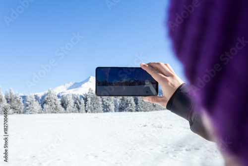 A person photographs a bright snowy mountain landscape with a smartphone, capturing the crisp winter scenery under a clear blue sky. Frosted trees and untouched snow create a serene outdoor atmosphere