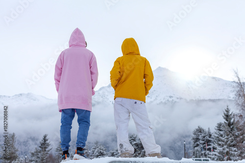 Two hooded figures in pink and yellow jackets stand on snowy ground, looking toward misty, snow-covered mountains beneath a pale winter sky.