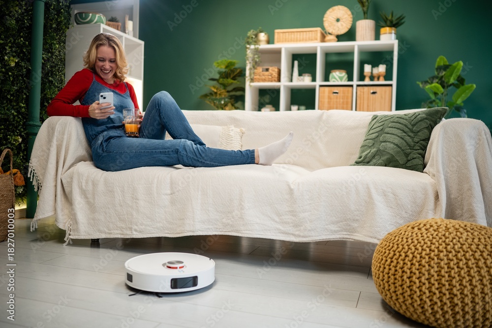 Obraz premium Woman relaxing with smartphone while robot vacuum cleaning