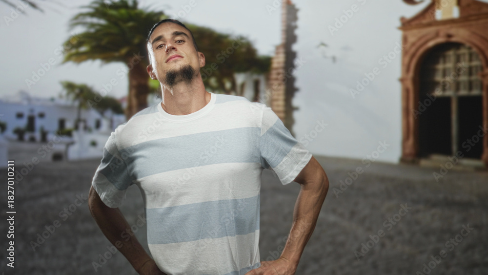 Fototapeta premium Man hands on hips posing in a sunlit street plaza beside an arched building, striped tshirt visible; confidence poise.