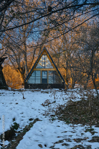 A-frame cabin in snowy forest at sunset, warm lights inside, winter retreat among leafless trees.