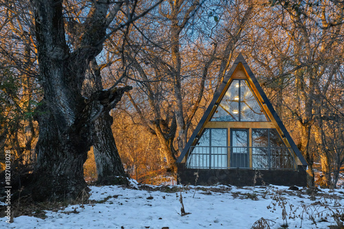 A-frame cabin in snowy forest at sunset, warm lights inside, winter retreat among leafless trees.