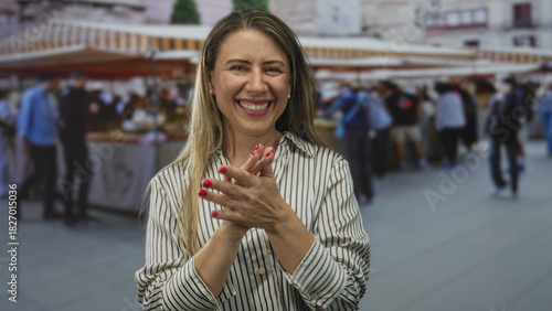 Woman clapping hands in bustling street market under striped awning beside vendor stall scene; celebration happiness.