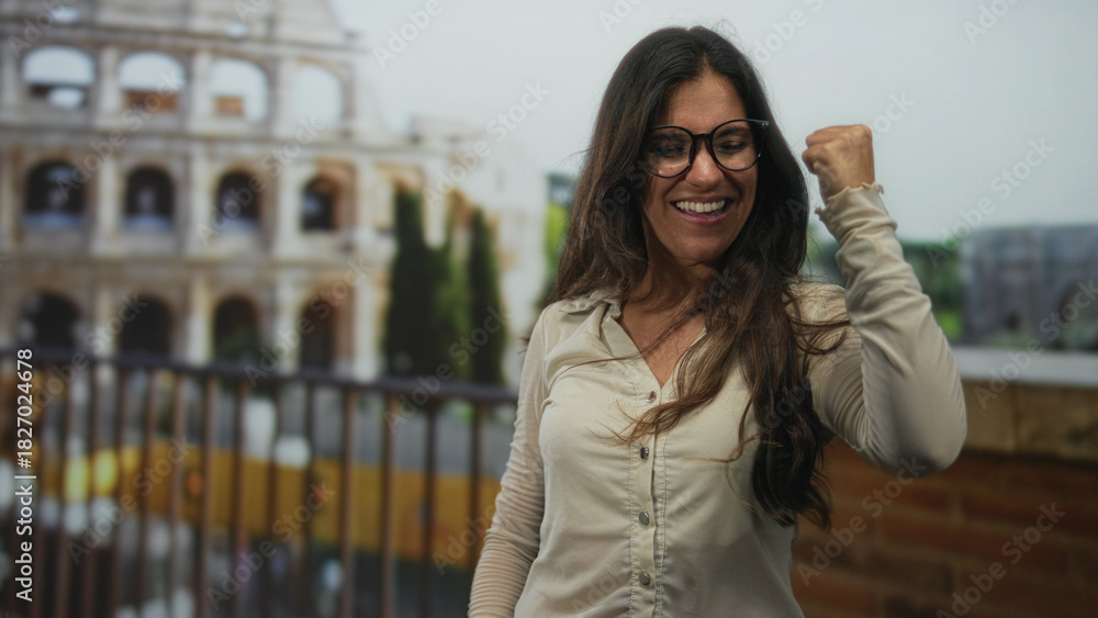Fototapeta premium Woman raising fists and smiling on coliseum terrace by a stone building railing, wearing glasses and casual shirt, celebrating a win; triumph resilience.