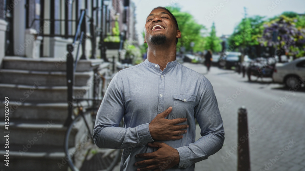 Fototapeta premium Young black man laughing with hand on chest and other hand on abdomen, wearing a button shirt by steps and bicycle on a street with parked cars; amusement.