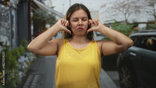 Woman in yellow tank top covering ears with fingers, eyes closed and grimacing on city street; annoyance sensory overload.