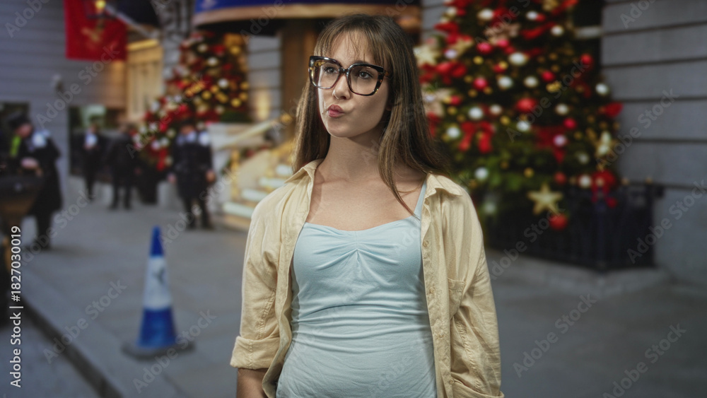 Fototapeta premium Woman wearing glasses and light blue tank top purses lips while standing by a decorated christmas tree on a city street; playful confidence.