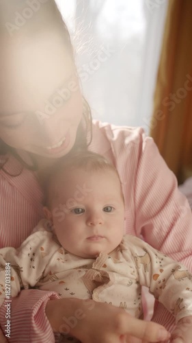 Affectionate young mother smiling warmly while cradling adorable baby in arms in room illuminated by morning sunlight. Vertical format video