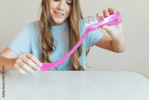 Happy girl pouring pink glitter slime from a bottle onto the table. Popular DIY activity for sensory fun, craft, and relaxation
