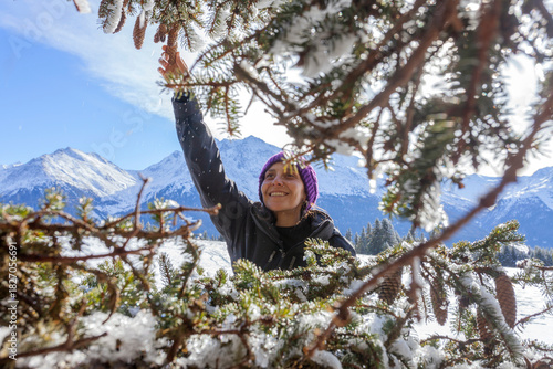 A woman in a purple beanie smiles as she reaches for pine cones on a snow-covered evergreen tree. Sunlight illuminates the winter forest and distant mountains, creating a lively and joyful outdoor mom