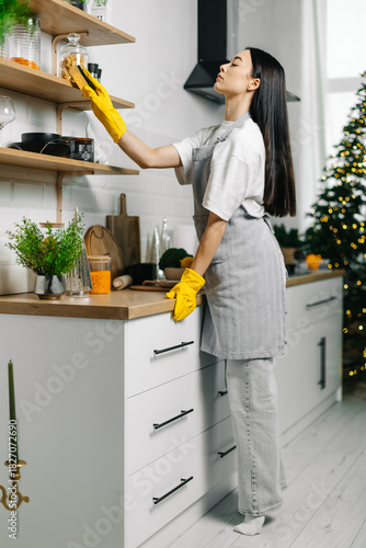 Full length woman cleaning kitchen shelves with yellow rubber gloves