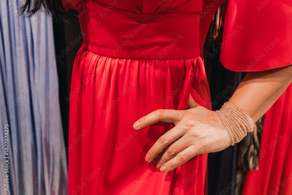 Fototapeta premium Elegant woman in a vibrant red dress poses confidently with hand on hip, showcasing the luxurious fabric and design details in a fashion retail environment