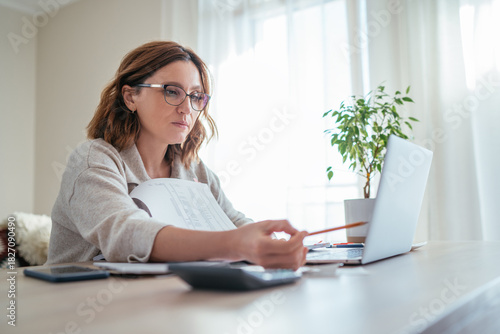 Beautiful middle-aged woman in glasses counting family business budget gazing at monthly bills and documents at home living room office. Small business home finances, money savings concept image