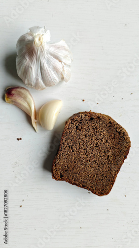 Slice of dark rye bread, head of garlic, and two garlic cloves on white wooden surface. Ingredients for healthy snack preparation for recipe blogs, cookbooks, health-conscious content. Rustic style