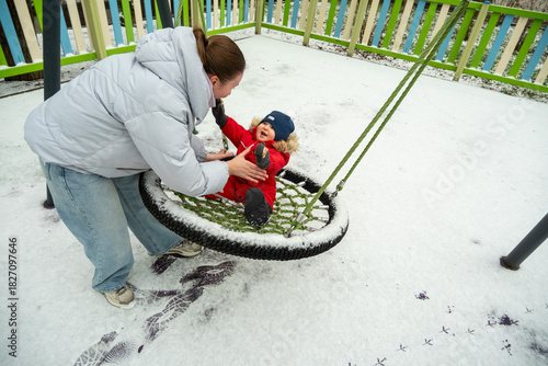 Child in winter clothing joyfully swings on a snow-covered playground swing while being gently pushed by a parent. The scene captures outdoor fun and bonding in a winter setting.