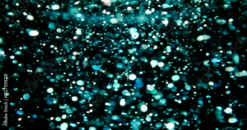 Particles float in water over a dark background, reflecting top light in green and blue hues, creating a glittering, halo-like, cosmic universe effect.