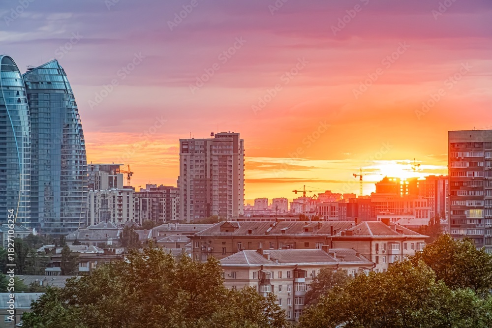 Fototapeta premium Vibrant Kyiv cityscape at sunset, with multiple high-rise buildings silhouetted against an orange and pink sky Some structures are illuminated, contrasting the urban landscape with a lush park in th