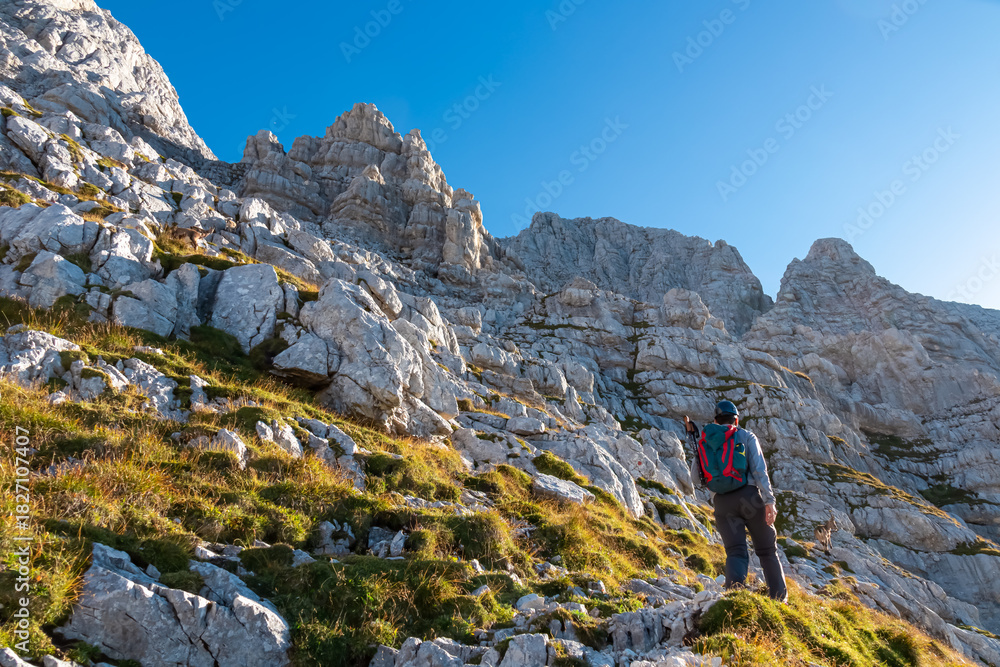 Naklejka premium A hiker equipped with a helmet and backpack ascends a steep rocky slope, gazing at the towering limestone cliffs of Jof di Montasio in the Julian Alps, Friuli Venezia Giulia, Italy, on a sunny day.