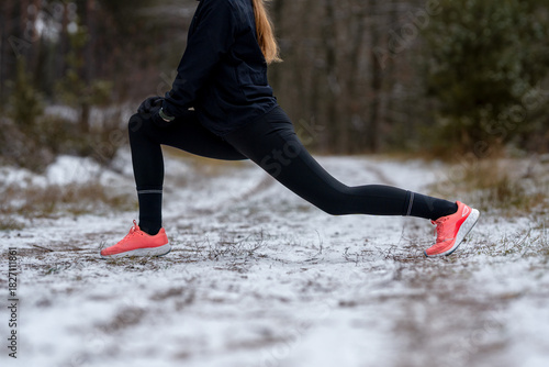 Focused on physical activity: Side view of a female runner performing a deep lunge to stretch the hamstring and hip flexor muscles on a snowy forest path.